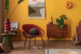 Aesthetic composition of warm and cozy living room interior with stylish kilim rug, wooden sideboard, plant, yellow wall, red armchair, brown sofa and personal accessories