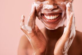 Person washing face with foamy cleanser on a pink background