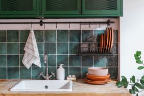 interior design in kitchen with water faucet, white sink, black metal drying rack with ceramic plates and glass, wooden countertop and green tile on wall