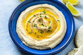 Vegan food: top view of a blue plate with hummus and olive oil shot on bluish tint table. Pita bread and some fresh vegetables on a cutting board like celery, baby carrots, radish and broccoli are around the plate and complete the composition