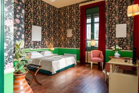 An inviting room with floral wallpaper and green wainscoting, featuring a retro style bed, elegant draperies, and classic furnishings, bathed in natural light from the balcony door.