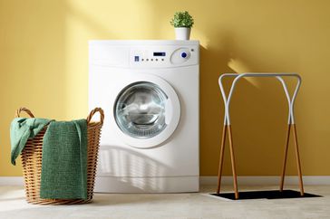 White modern washing machine, rattan basket with towel and cloth rack on tile floor in empty vibrant yellow wall laundry room with beautiful sunlight from window for washing and cleaning product display