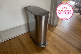 The Simplehuman 12-Gallon Slim Kitchen Trash Can on a wood floor next to a wall.