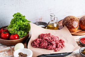 Organic raw ground beef on butcher paper, burger ingredients on white wood rustic table, preparing food. Table with tomatoes, salad, onion, oil, hamburger buns, pickles, and spices