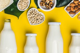 Row of Non-Dairy Milks in Glass Bottles With Bowls of Nuts, Rice, and Oat Above Them, yellow background